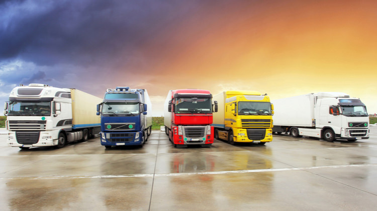 Five colourful lorries parked in a row on a wet concrete surface, with a dramatic sunset sky in the background.